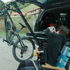 Person sitting next to bike rack with trunk open. Bike rack is tilted for trunk access. 