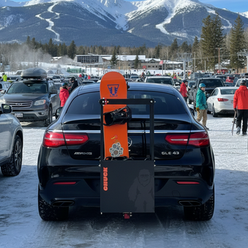 Car with an orange snowboard attached to a chuck bucket hitch mounted ski rack in a snowy parking lot with mountains in the background.