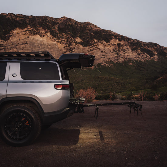 A silver off-road SUV parked outdoors at dusk with its rear hatch open, towing a black trailer. A rocky mountain rises in the background, and the vehicle is shown in a real outdoor environment.