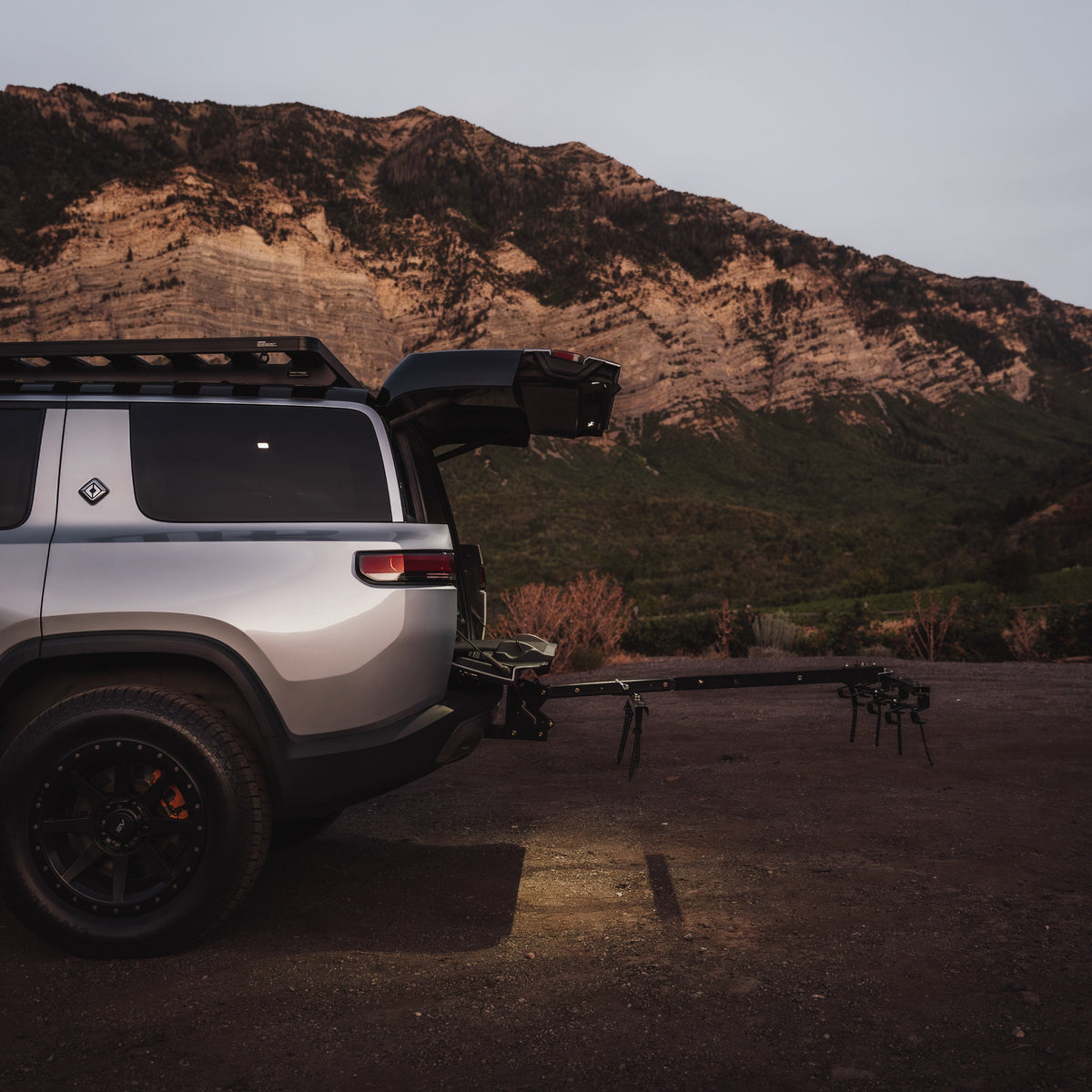 A silver off-road SUV parked outdoors at dusk with its rear hatch open, towing a black trailer. A rocky mountain rises in the background, and the vehicle is shown in a real outdoor environment.